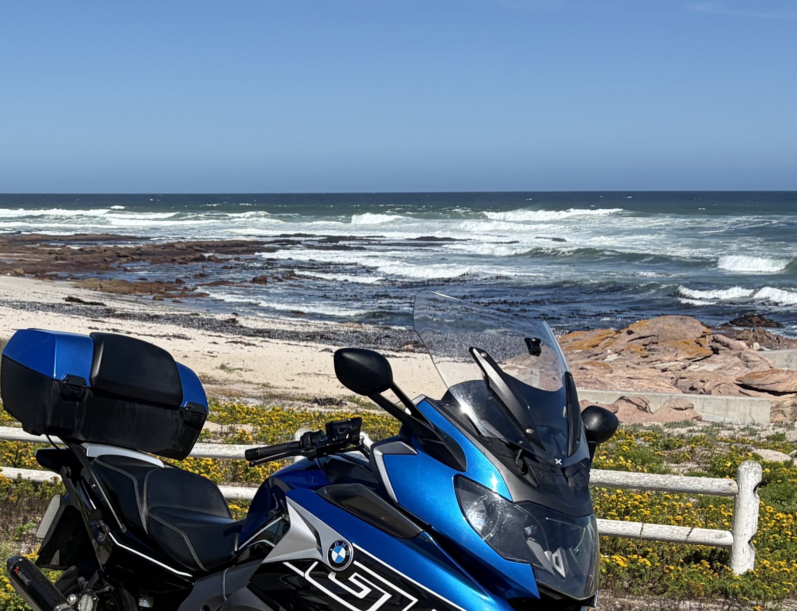 The same BMW K1600 GT parked above a rocky stretch of South African coastline, with breaking surf beyond.
