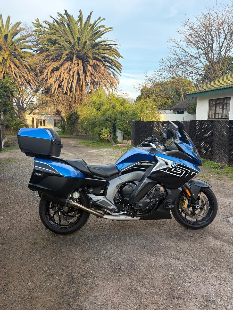 A blue BMW K1600 GT in side profile, with panniers and a top box fitted, parked on a quiet lane lined with palms.