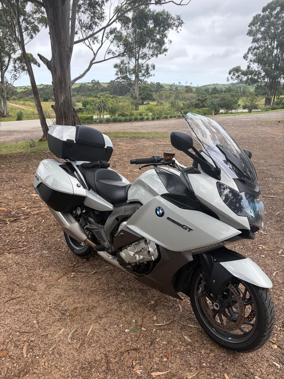 A white BMW K1600 GT with panniers and a top box, seen from the front three-quarter, parked under gum trees.
